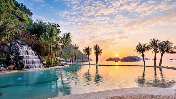 Un área de piscina tranquila con una cascada, rodeada de palmeras y vistas al atardecer.