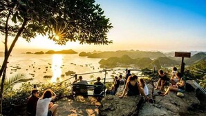 Un grupo de personas sentadas sobre rocas, contemplando un cuerpo de agua con el atardecer de fondo.