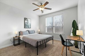 Ein Schlafzimmer mit einem Bett, zwei Nachttischen, einem Schreibtisch, einem Sessel, einem Deckenventilator, einem Fenster mit Jalousien und einem an der Wand hängenden Bild.