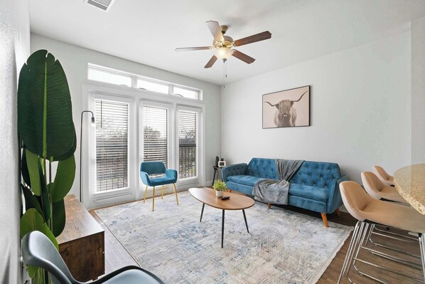 Ein modernes Wohnzimmer mit einem blauen Sofa, einem runden Couchtisch, einem Deckenventilator und großen Fenstern.