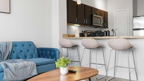 Uma sala de estar moderna com um sofá azul e uma mesa de centro de madeira. Há três banquetas e uma pequena mesa com uma planta e um controle remoto. A cozinha ao fundo tem armários escuros, um micro-ondas e uma geladeira.