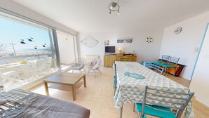 Ein Zimmer mit großem Fenster, einem Esstisch, Stühlen, einer Couch, einem Fernseher und einer kleinen Küchenzeile.