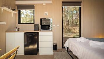 Quarto compacto com cozinha compacta, uma cama e toalhas dobradas, com vista para uma floresta através de uma grande janela.