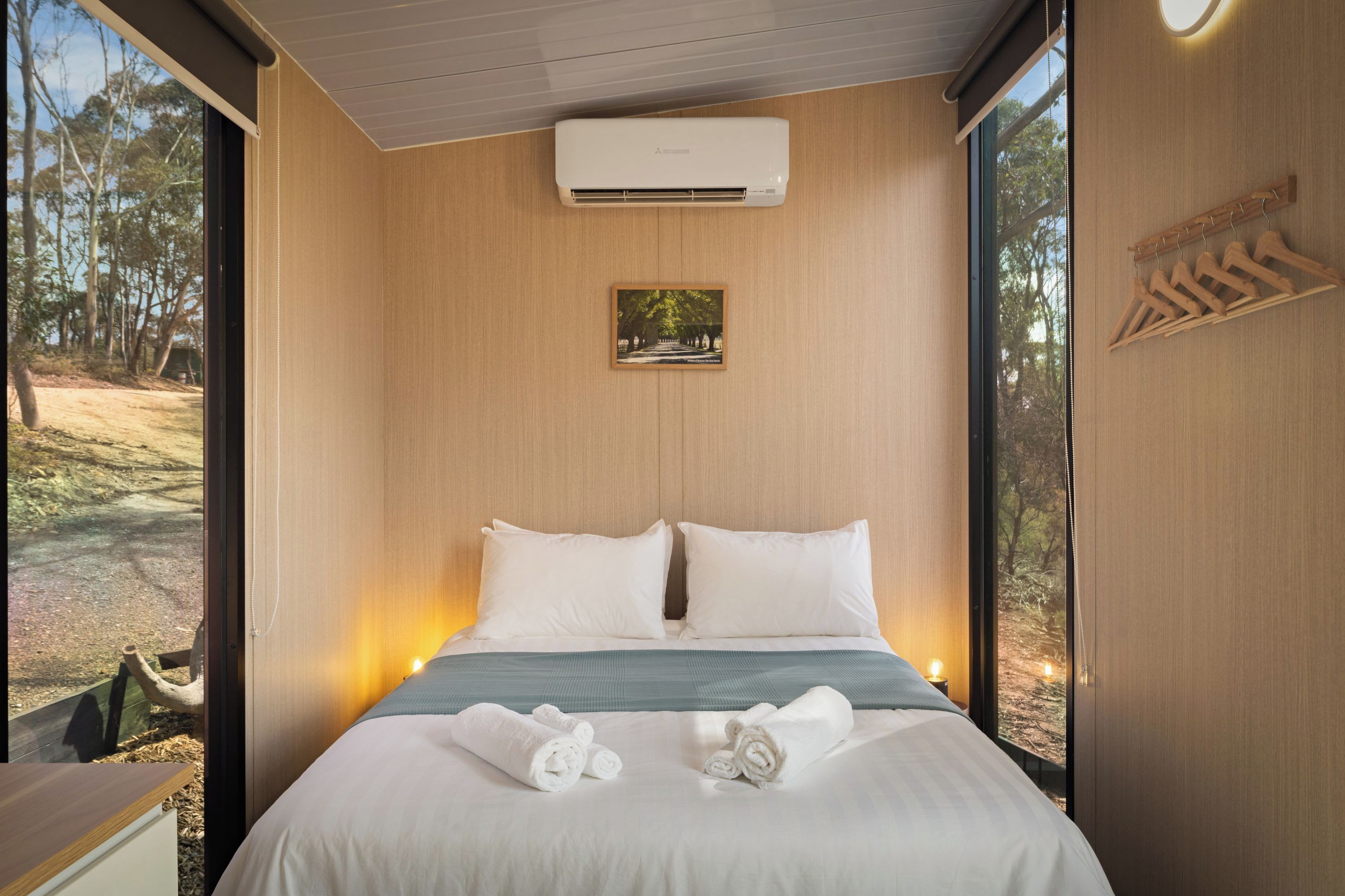 Een moderne slaapkamer met een groot bed, een vastgemaakt airco-apparaat, een ingelijst kunstwerk en houten kledinghangers aan de muur.
