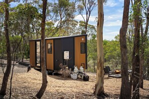 A small wooden cabin with a black door, surrounded by trees. It has a fire pit and outdoor seating. - Eucalypt Escape by Tiny Away (Coimadai)