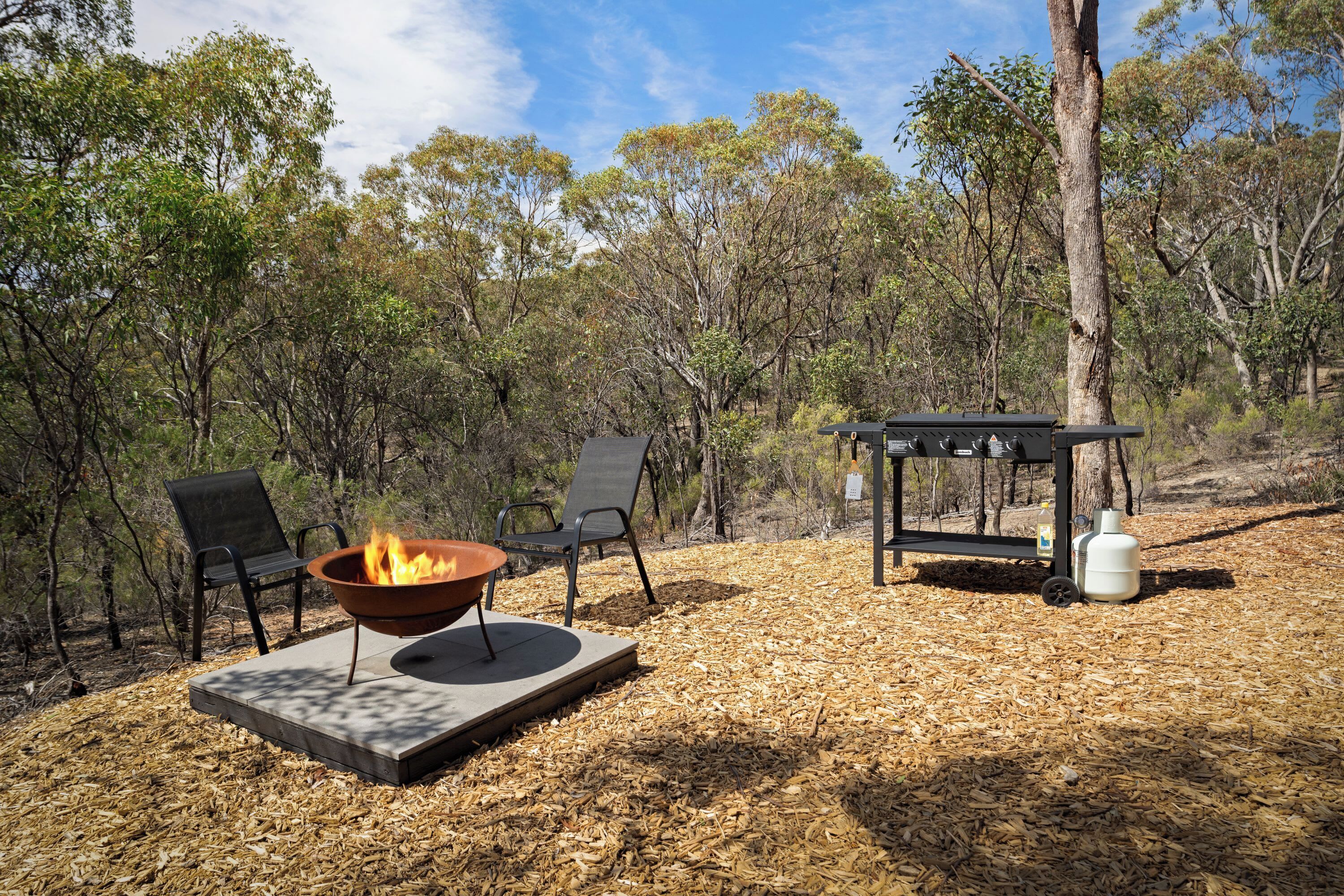 A fire pit with two chairs, a table, and a gas cylinder in a forested area.