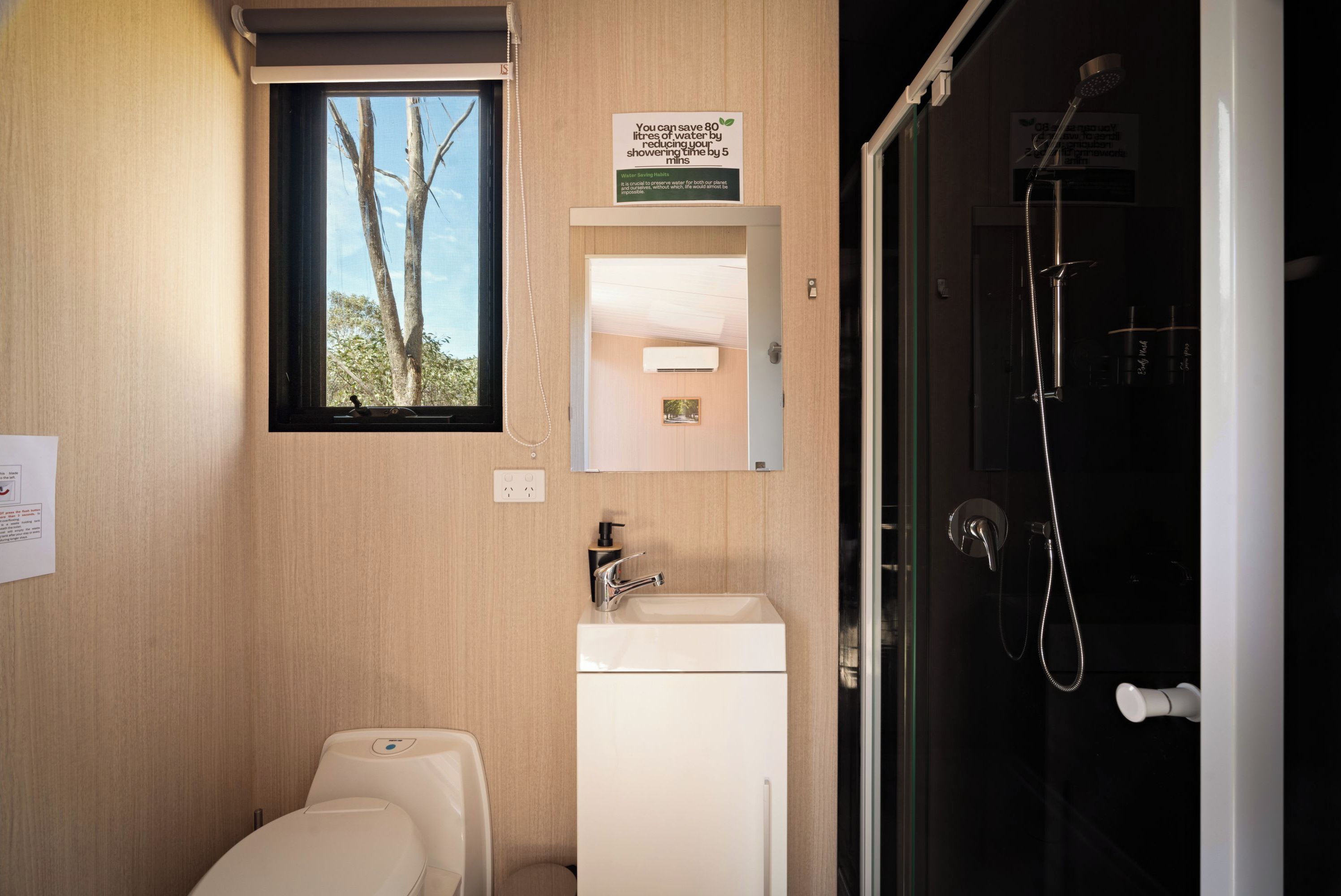 Ein kleines Badezimmer mit Toilette, Waschbecken und Dusche. Ein Fenster sorgt für natürliches Licht, und ein Schild über dem Spiegel gibt Tipps zum Wassersparen.
