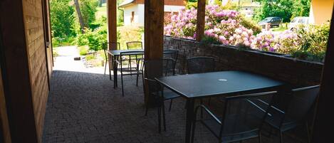 Un patio exterior con una mesa y sillas, piso de piedra y vista a un jardín con flores y una casa al fondo.