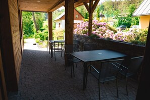 An outdoor patio with a table and chairs, a stone floor, and a view of a garden with flowers and a house in the background. - Villa in Jeseniky With Sauna & Mountain Views (Bela pod Pradedem)
