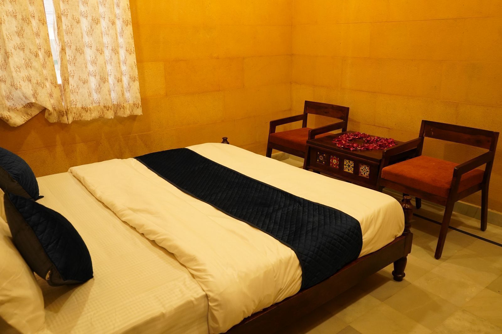 A room with a bed, two chairs, a small table with a red cloth, and a window with patterned curtains.