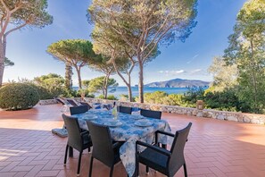 An outdoor dining area with a table set for six, surrounded by stone walls and trees, overlooking a scenic coastal view. - Villa with private access to the beach (Capoliveri)