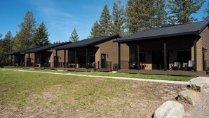 Un hilera de cabañas modernas con zonas de estar al aire libre, rodeadas por un campo de césped y altos árboles.