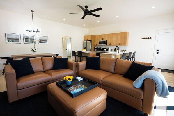 A modern living room with a brown leather sectional sofa, a coffee table with a book and flowers, a ceiling fan, and a kitchen in the background.