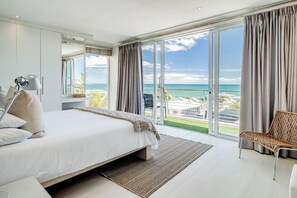 Enjoy ocean views from the large bed in this serene bedroom, featuring a window with curtains, a bedside lamp, and a wicker chair.