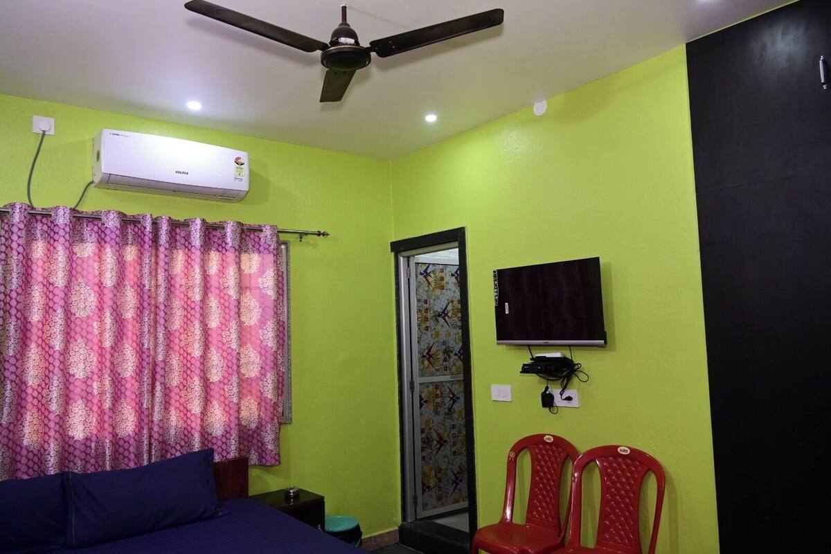 A room with a bed, a ceiling fan, an air conditioner, a TV, and two red chairs.
