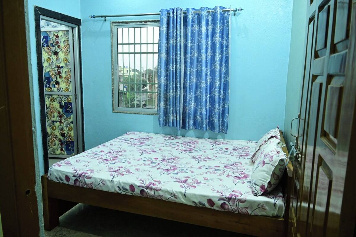 A small, single-bed room with a floral bedspread, a window with a metal grill, and a blue curtain.