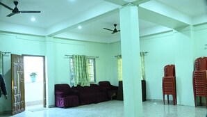 Une pièce spacieuse avec un plafond haut, des ventilateurs de plafond, un coin salon avec un canapé et des fauteuils, et une porte donnant sur l’extérieur.