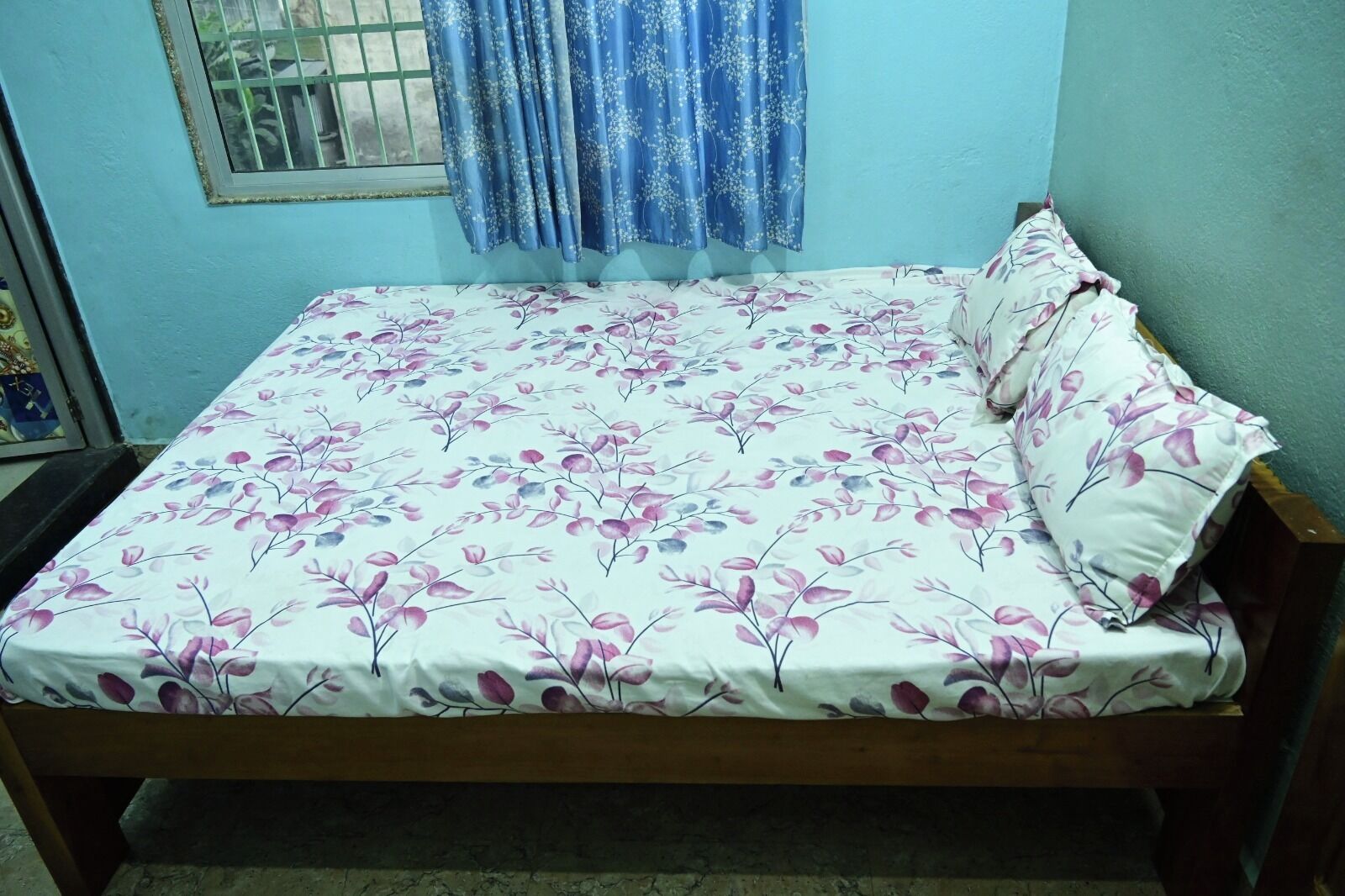 Una cama con colcha y almohadas estampadas con flores, una ventana con rejas y una cortina azul.