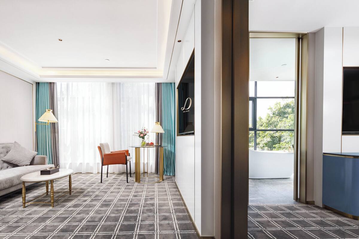 Een moderne hotelkamer met een bank, een klein tafeltje met een lamp, een stoel, en een groot raam met uitzicht op het omliggende landschap.