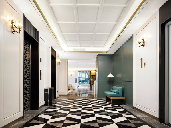 A hotel lobby with a geometric patterned floor, a seating area with a teal sofa, and a view of the cityscape through the glass doors.