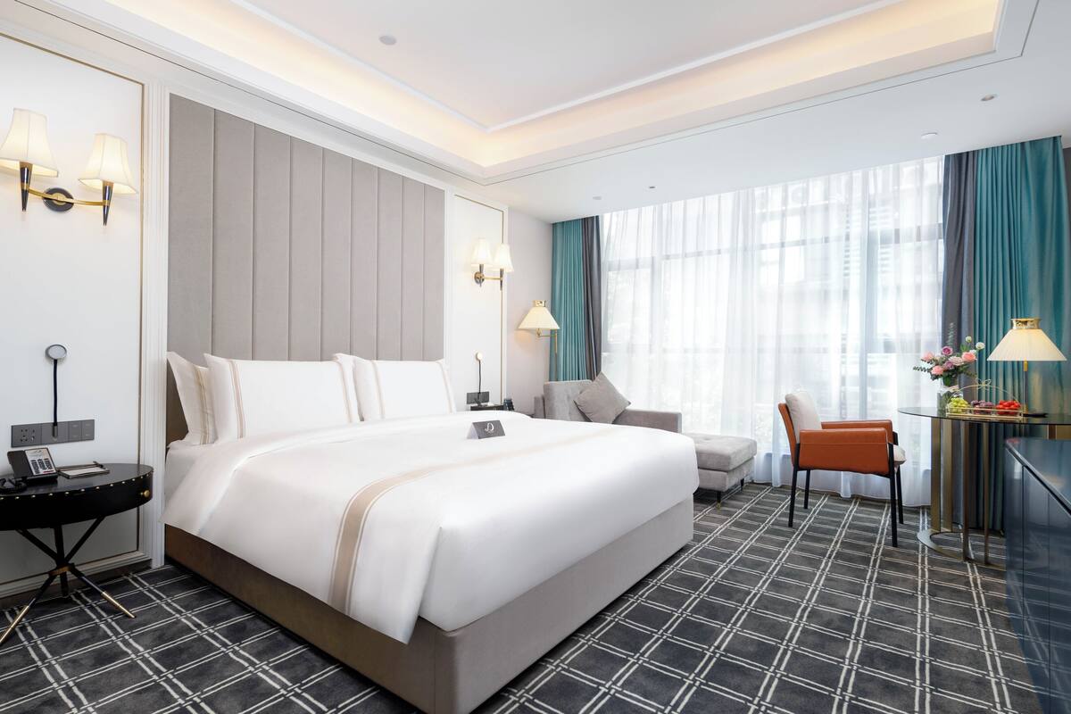 Hotelkamer met een groot bed, nachtkastjes, een bureau met lamp, een stoel en uitzicht op de stad door grote ramen.