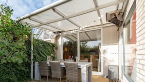 En udendørs terrasse med et bord og stole, en glasdør der fører til en have, og en hvid markise.