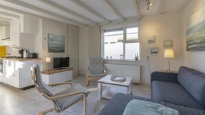 Een moderne woonkamer met een bank, twee fauteuils, een salontafel en een televisie. Er is een keuken met een aanrecht en apparatuur. De kamer heeft grote ramen en een plafond met zichtbare balken.