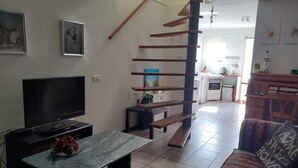 Un salon avec un téléviseur, une table basse en marbre, un canapé agrémenté de coussins à motifs, un escalier en bois et, au fond, une cuisine.