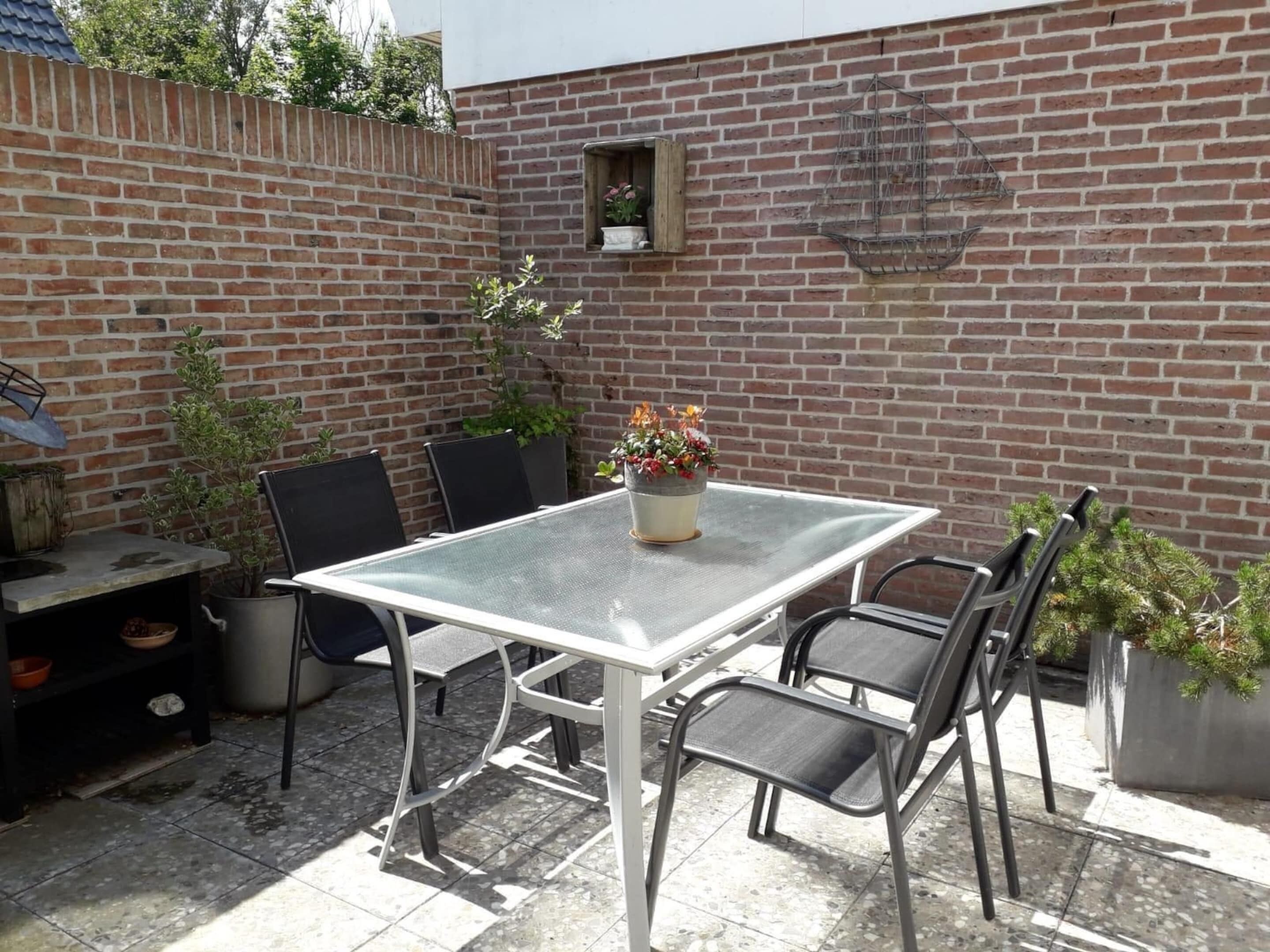 Une terrasse avec une table en verre et des chaises en métal, entourée de plantes en pot et d’un mur en briques.
