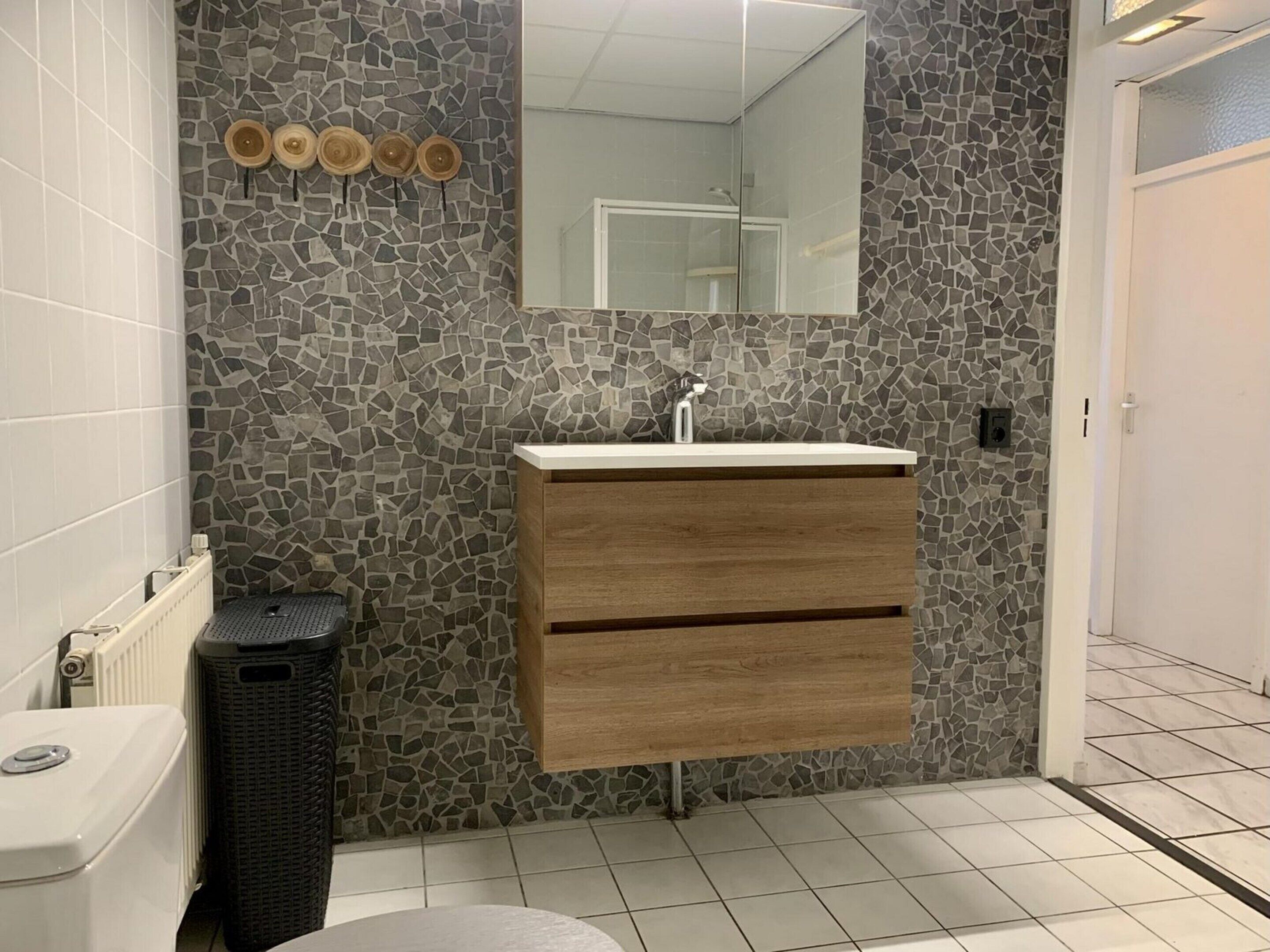 Une salle de bain avec un mur décoré de mosaïques, un meuble-lavabo en bois, un lavabo blanc et des toilettes.