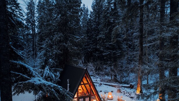Un chalet doté d’un toit triangulaire, entouré d’arbres couverts de neige, et un bain à remous sur la terrasse.