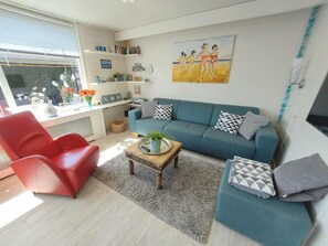 Ein Wohnzimmer mit einem blauen Sofa, einem roten Sessel und einem Couchtisch. An der Wand hängt ein Gemälde und es gibt ein Fenster mit Jalousien.