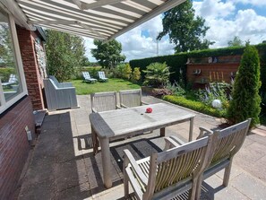 Une terrasse couverte avec une table et des chaises, un mur en briques et un jardin avec une pelouse, des chaises et des plantes.