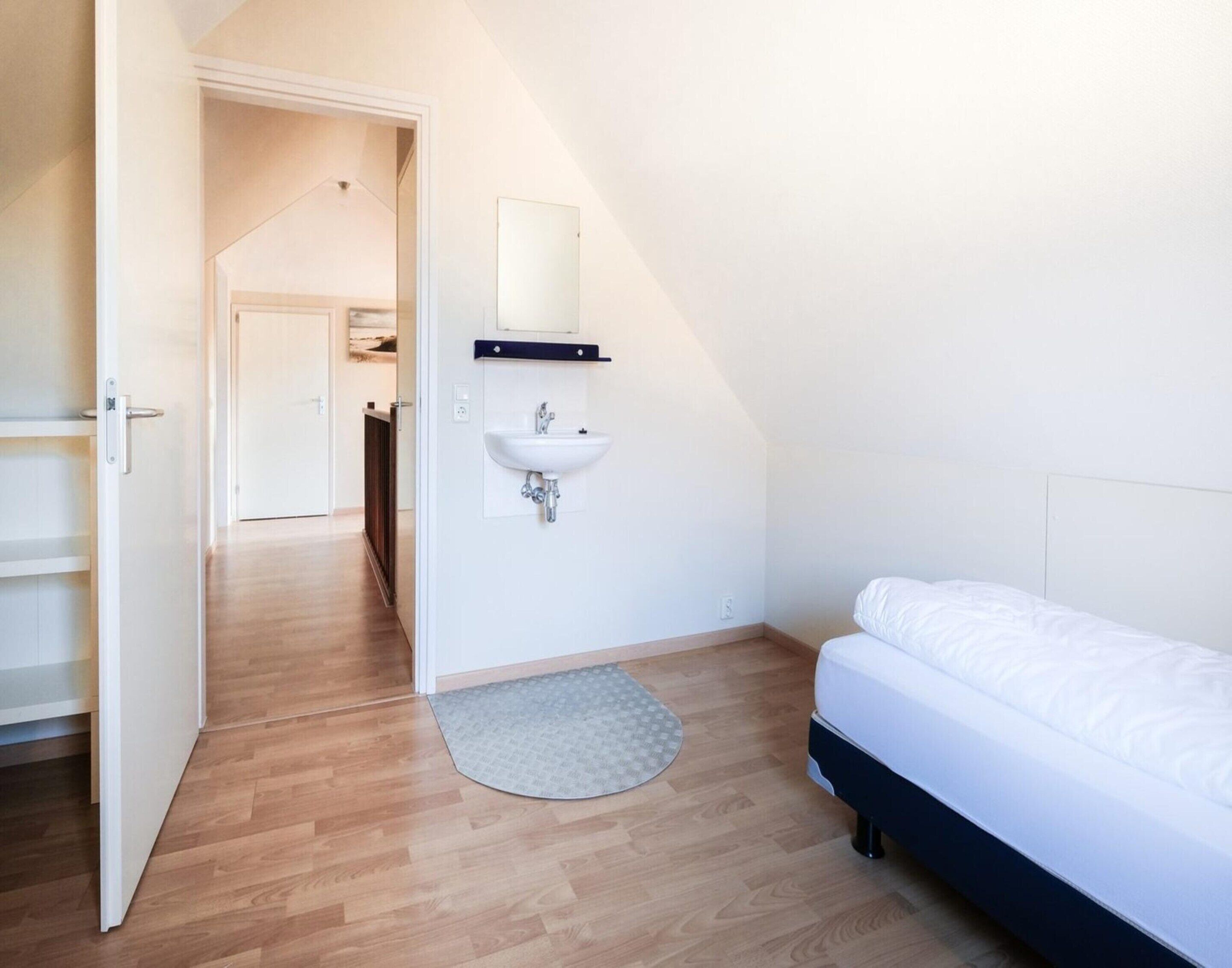 Een slaapkamer met een bed, een wit wandmodel wastafel, een houten vloer en een deur die naar een andere kamer leidt.