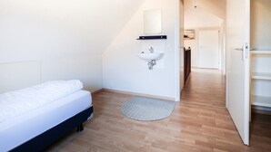 Un dormitorio con una cama, una mesita de noche, un lavabo blanco instalado en la pared y una puerta que da a otra habitación.