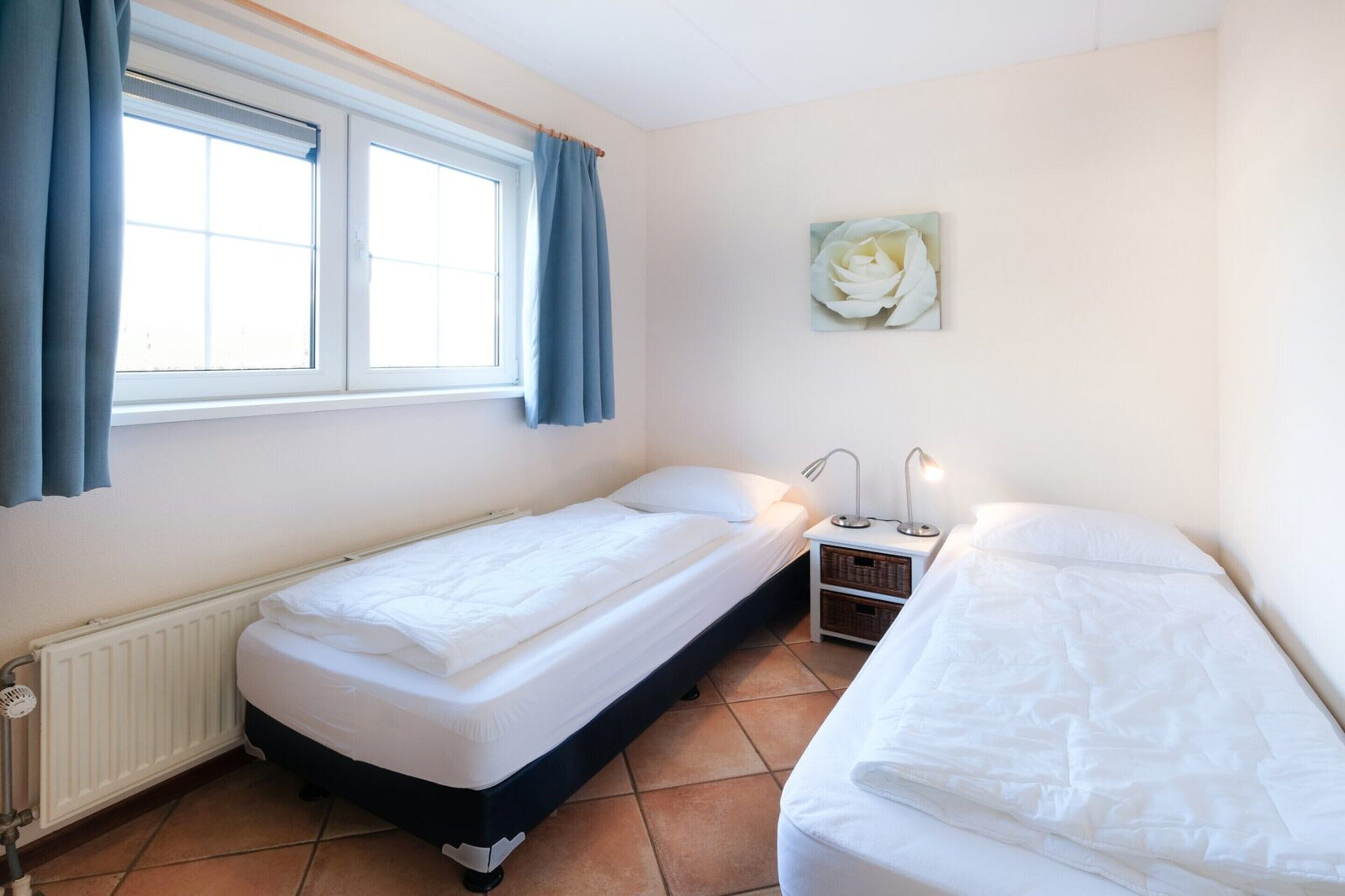 Une petite chambre propre, avec deux lits simples, une fenêtre avec des rideaux, un radiateur et une lampe fixée au mur.