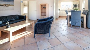Una sala moderna con un sofá, una mesa de centro y sillones de mimbre. Se observa una zona de cocina con una mesa de comedor y sillas.