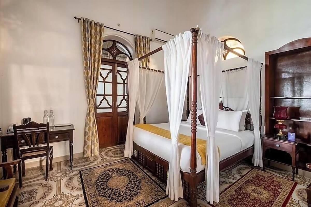 A four-poster canopy bed with a wooden frame and white bedding, a desk with two bottles, and a patterned rug.
