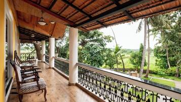 A covered balcony with wooden flooring, patterned chairs, and a view of a lush green landscape.