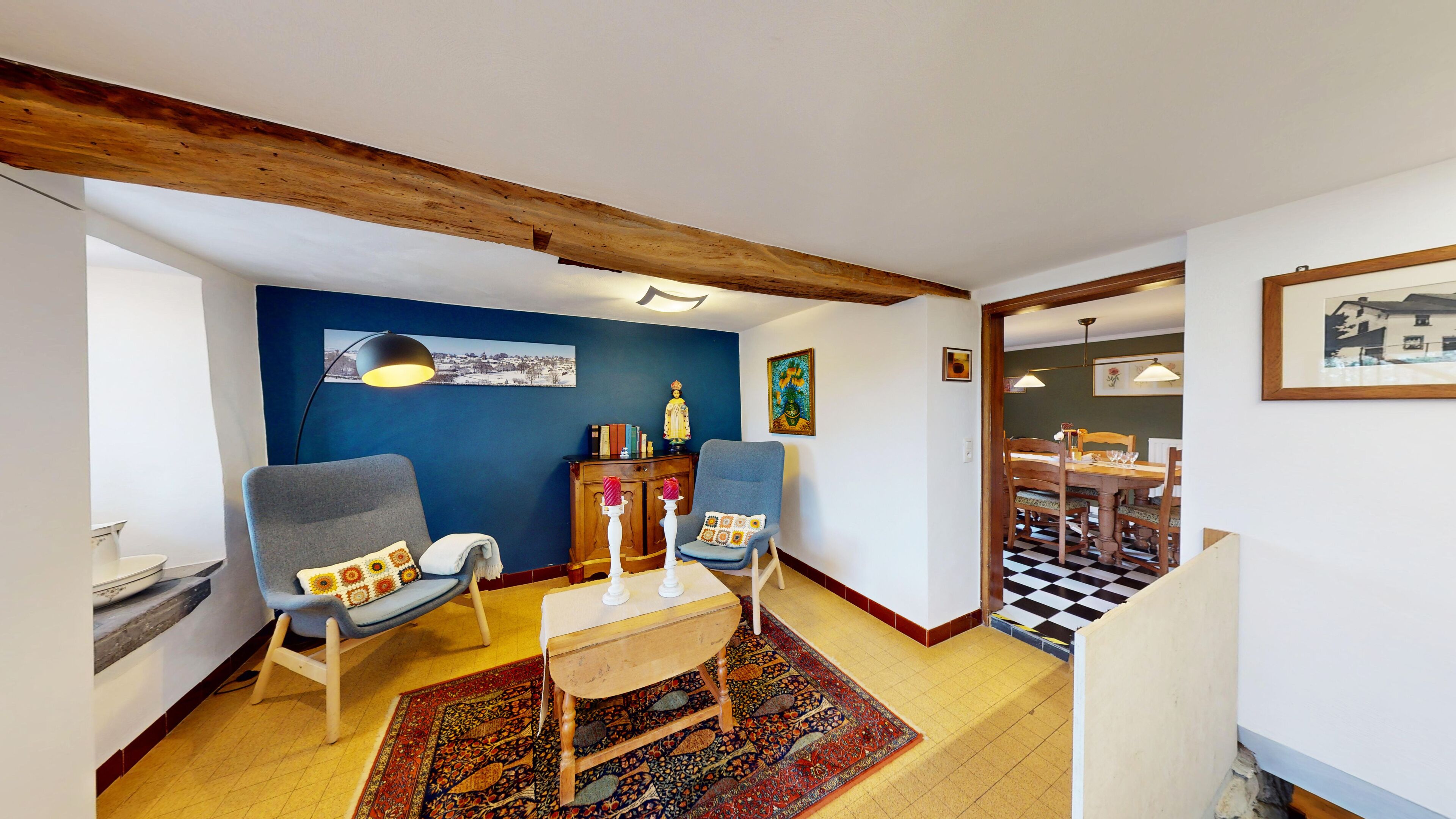 A cozy living room with a blue accent wall, a wooden ceiling beam, a dining area with a checkered floor, and a view into another room.