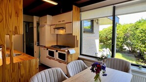 A compact kitchen with built-in appliances, a dining area with a wooden table and chairs, and a large window offering a view of greenery.