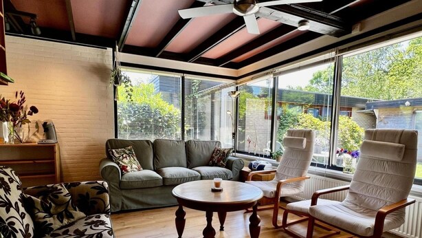 A sunroom with a ceiling fan, a grey sofa, a round wooden coffee table, and a white armchair.