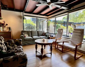 Une véranda avec un ventilateur de plafond, un canapé gris, une table basse ronde en bois et un fauteuil blanc.