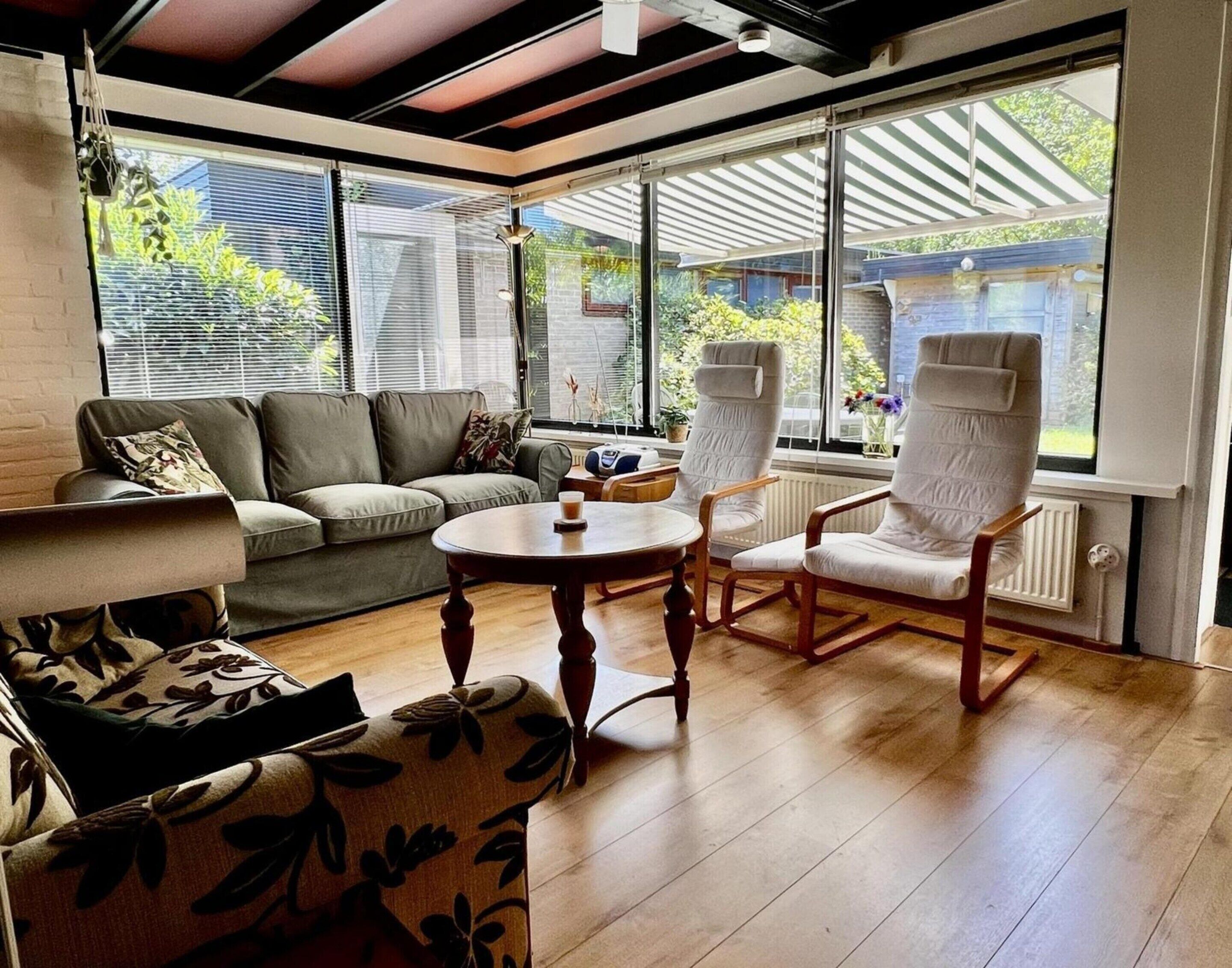 Un salon moderne comprenant un canapé, deux fauteuils, une table basse et de grandes fenêtres équipées de stores.
