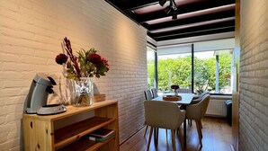 A modern dining area with a wooden table and chairs, a vase with flowers, and a coffee machine on a wooden shelf.