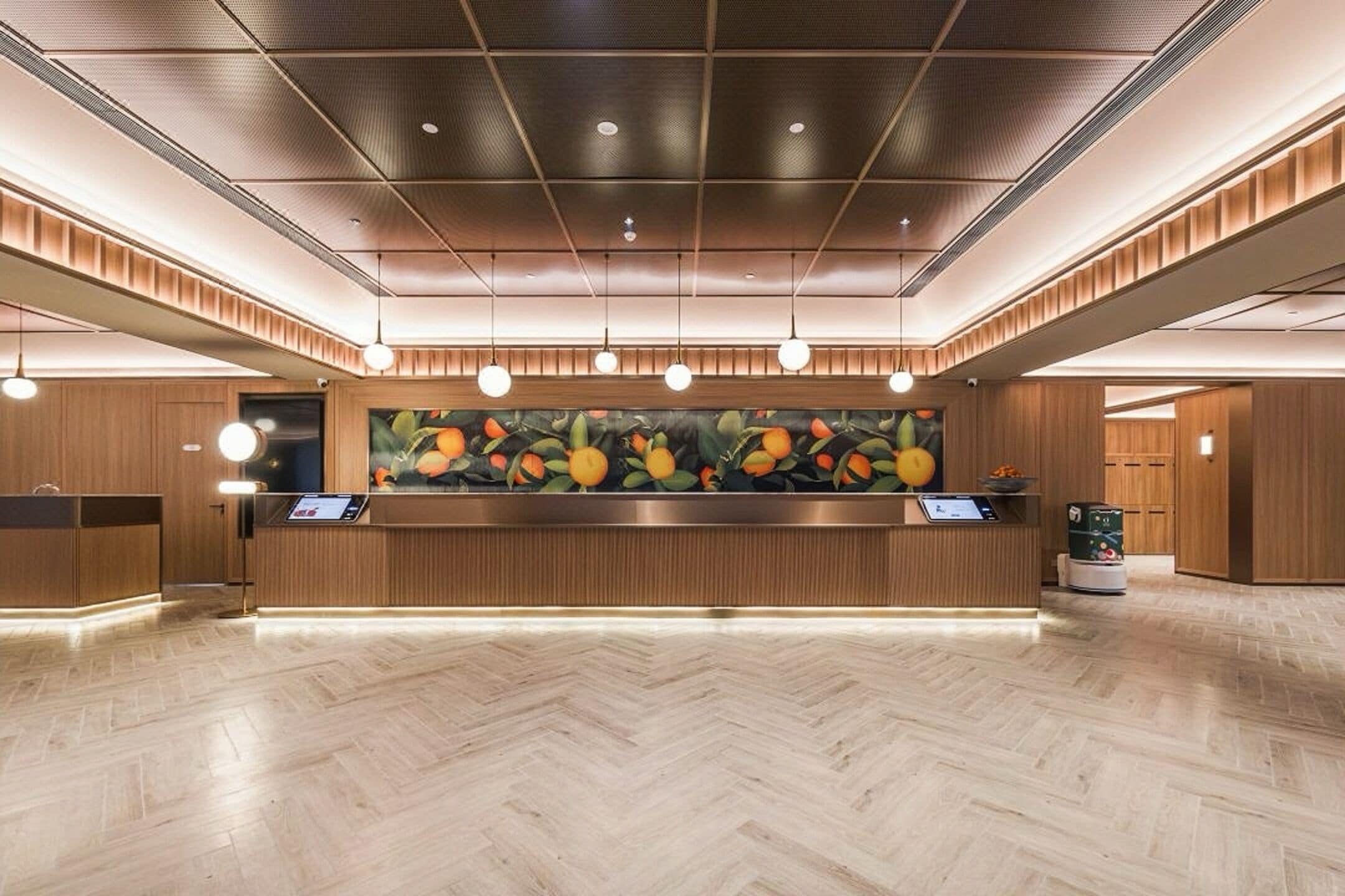 Un área de recepción con piso de madera, un mostrador central con pantalla digital y un cuadro de gran tamaño con motivos de frutas sobre el mostrador.