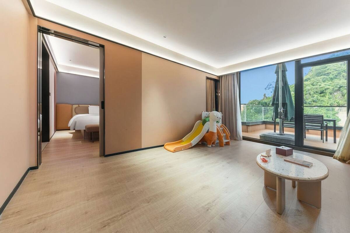 Habitación de hotel con piso de madera, una cama, una mesita, un tobogán infantil y un balcón con mobiliario exterior.
