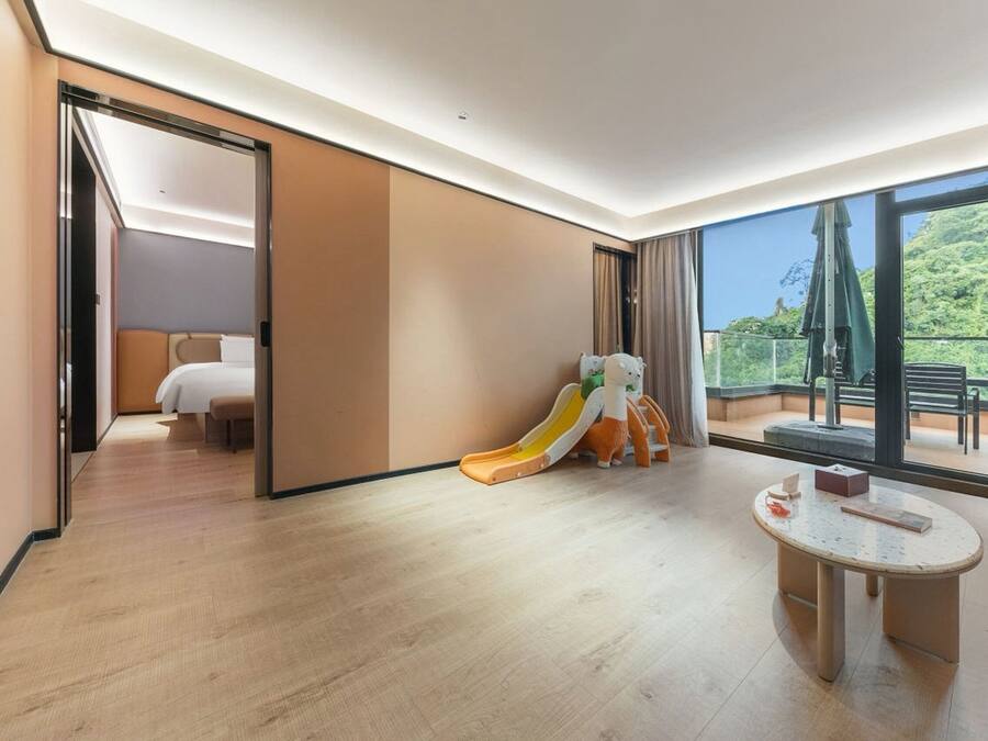 A hotel room with a wooden floor, a bed, a small table, a children's slide, and a balcony with outdoor furniture.
