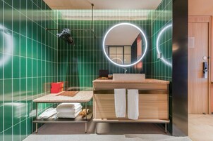 Un baño moderno con paredes de azulejos verdes, un mueble de tocador de madera con lavamanos, un espejo con luz circular y un perchero para toallas.