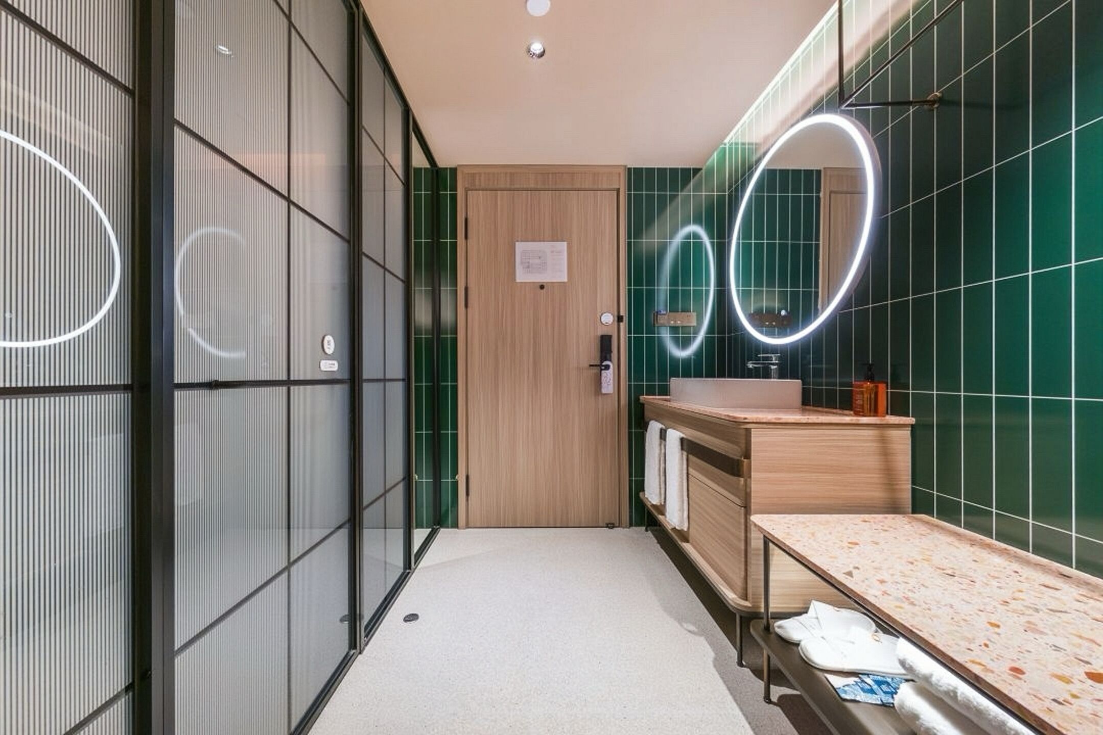 A modern bathroom with a wooden door, a sink with a mirror, and green tiled walls.